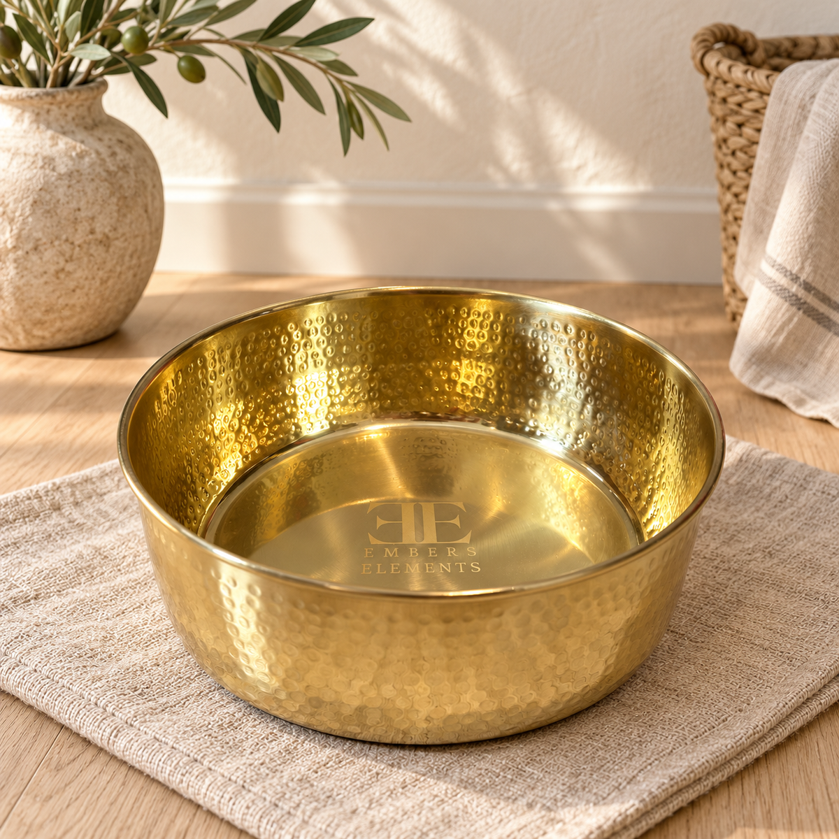 AURORA hand-hammered brass pet food bowl on a woven mat beside an olive branch and towel basket