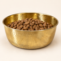 AURORA hand-hammered brass pet food bowl filled with dry kibble, studio shot on a cream background