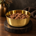 DUNE smooth-finish brass pet food bowl filled with wet meat-chunk food, moody studio setting with a brass vase